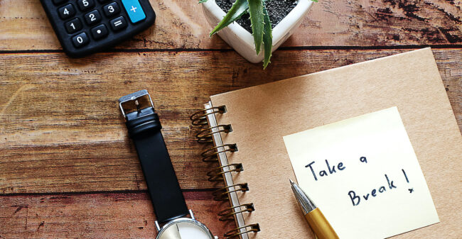 Wood Desktop work space have brown glasses,calculator,pot plant ,black watch,pen,notebook,and note pad with “Take a break”message.For Work life balance concept,Happy work place,Have copy space. work place stress
